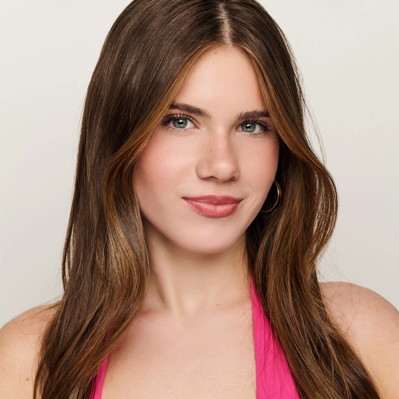 A young woman with long, straight brown hair and blue eyes wears a hot pink halter top and gold hoop earrings. She has a natural makeup look with rosy cheeks and soft pink lipstick. The background is plain and light-colored, creating a simple yet striking portrait reminiscent of Jessica Evgenikos’ effortless style.