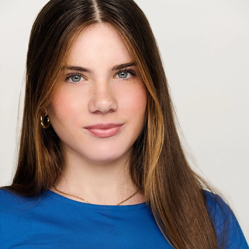 Jessica Evgenikos, a young woman with fair skin, long straight brown hair, and blue eyes, faces the camera. She wears a bright blue top, gold hoop earrings, and a thin necklace. The light gray background complements her slight smile.