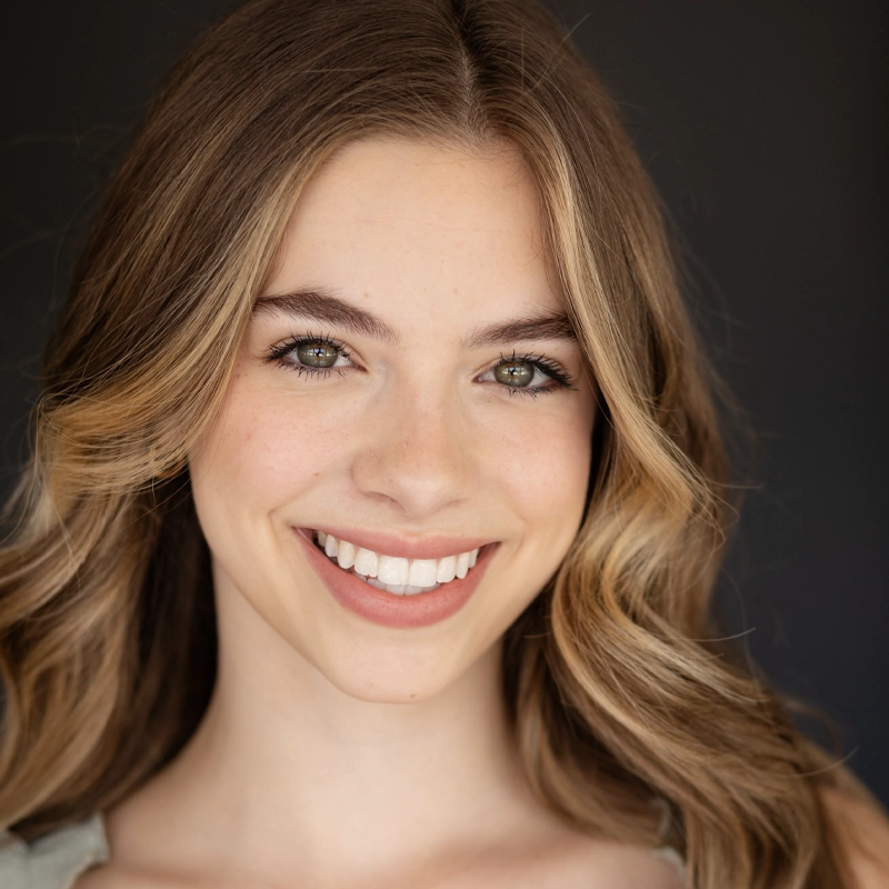 Savannah Fouchi, a young woman with light skin and long, wavy, light brown hair, smiles warmly. She has green eyes, natural makeup, and stands against a dark, blurred background in a light-colored top, looking directly at the camera.
