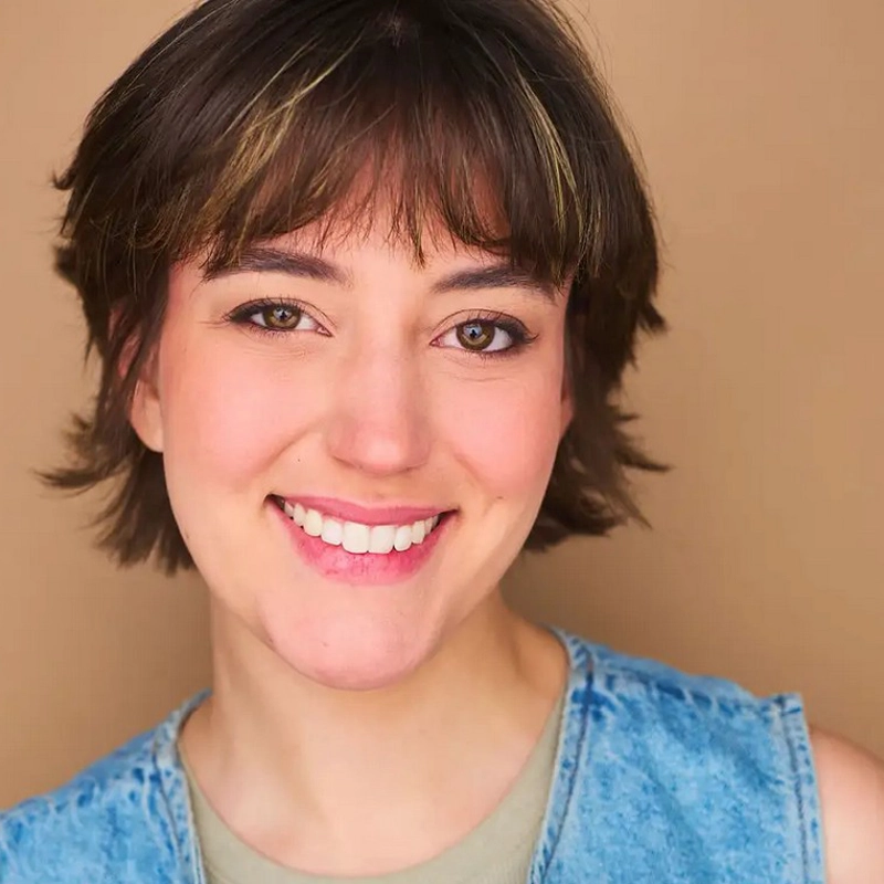 Sydney Freihofer, a young person with short, tousled brown hair and bangs, smiles warmly. With fair skin and brown eyes, they wear light makeup and a sleeveless light blue denim top over a beige shirt against a plain tan background.