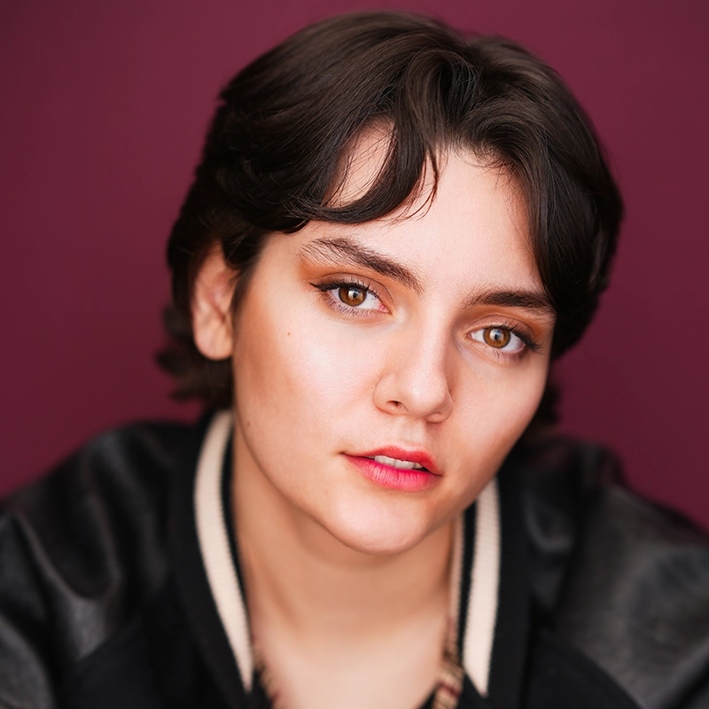 A young person with short brown hair and light skin looks directly at the camera. With brown eyes, subtle makeup, and natural pink lips, Mickey Wirtz wears a dark jacket with a light collar against a solid burgundy background.