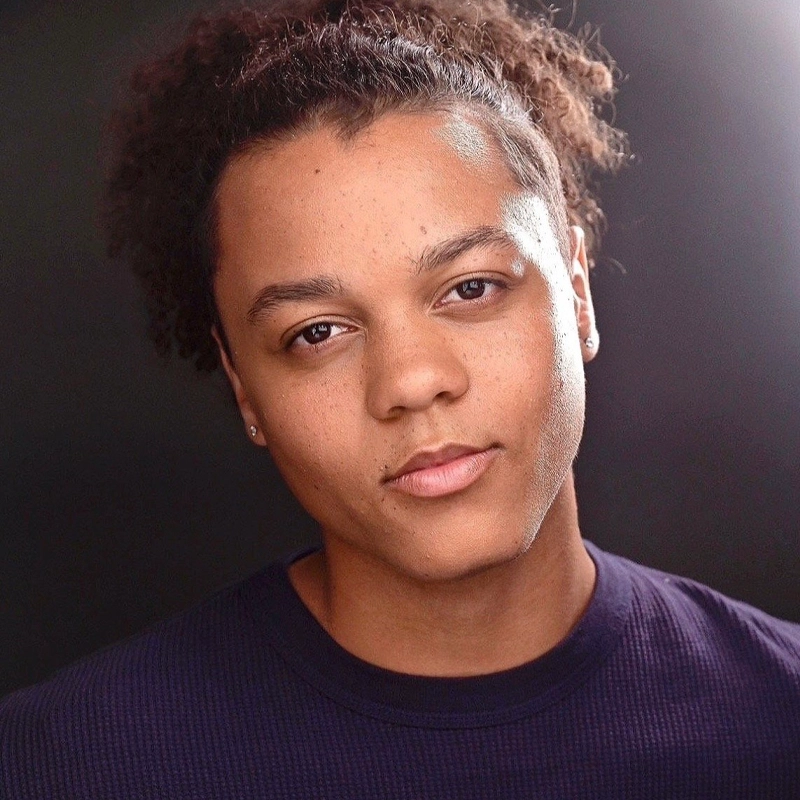 Sebastian Gates-Graceson, a young person with medium brown skin, curly dark brown hair pulled back, and small earrings, looks directly at the camera with a calm, neutral expression. They wear a dark ribbed shirt under soft, even lighting against a dark background.