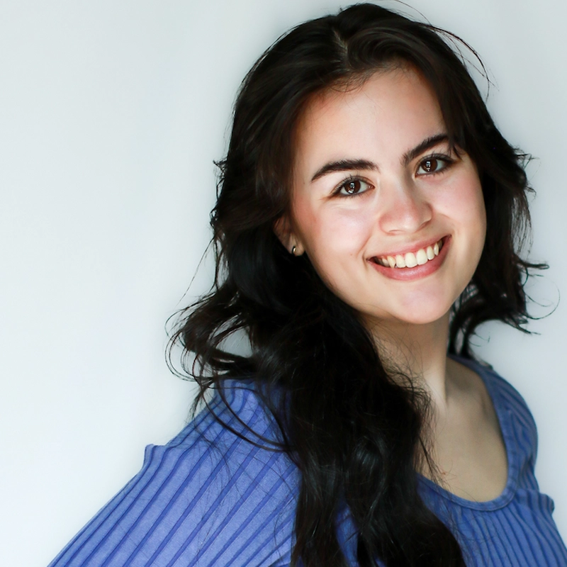 MacKenzie Goode, a young woman with long, wavy dark hair, smiles warmly at the camera. She wears a ribbed, blue top and stands against a plain, light background, her head slightly tilted and fair skin glowing with natural makeup.