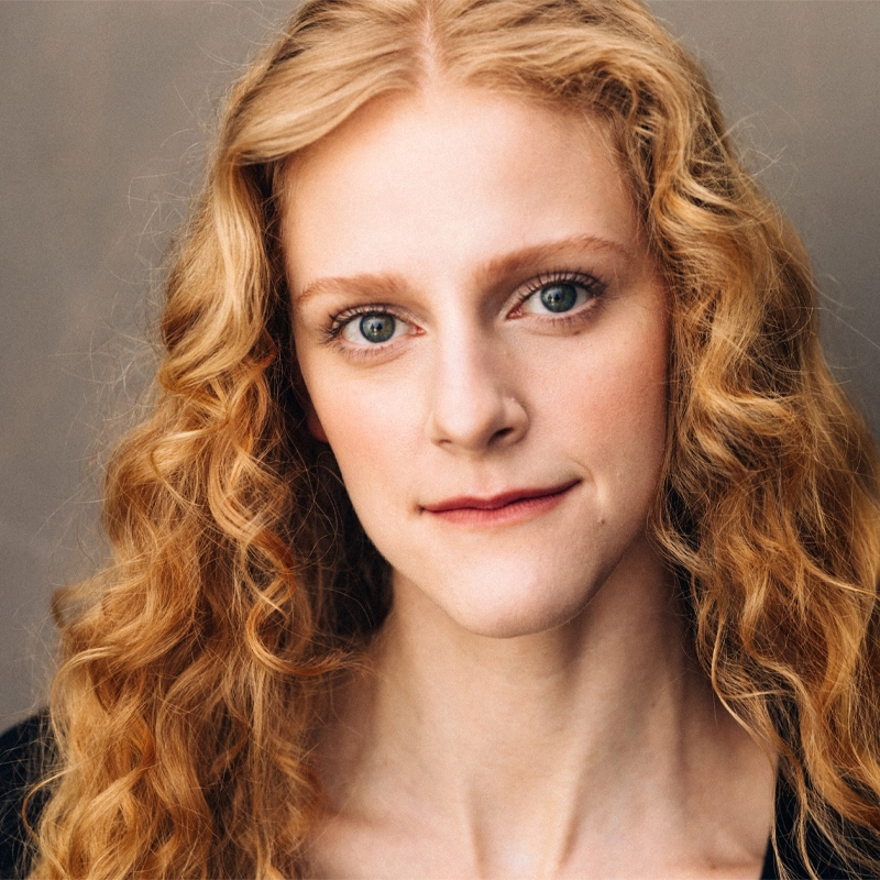 Katie Griffith, a young woman with long, curly strawberry blonde hair and fair skin, looks directly at the camera with a neutral expression. Her blue eyes and natural makeup stand out against her dark top and the soft, muted background.