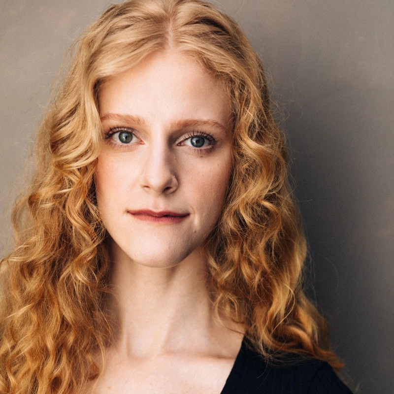 Katie Griffith, with long, curly blonde hair and fair skin, looks directly at the camera. She has blue eyes, subtle makeup, and wears a black top. The softly blurred neutral background draws focus to her face.