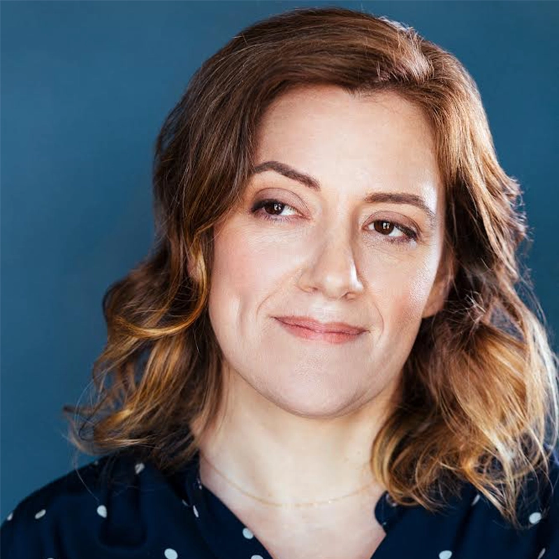 A woman with wavy, shoulder-length light brown hair and fair skin looks slightly to her left, smiling subtly. She wears a dark blue top with white polka dots against a solid blue background, reminiscent of Rickey Tripp’s stylish flair.