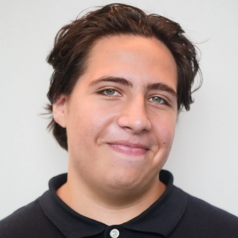 Michael Hernandez, a young person with medium-length brown hair and light skin, smiles softly at the camera. He is wearing a dark polo shirt and stands in front of a plain, light-colored background.