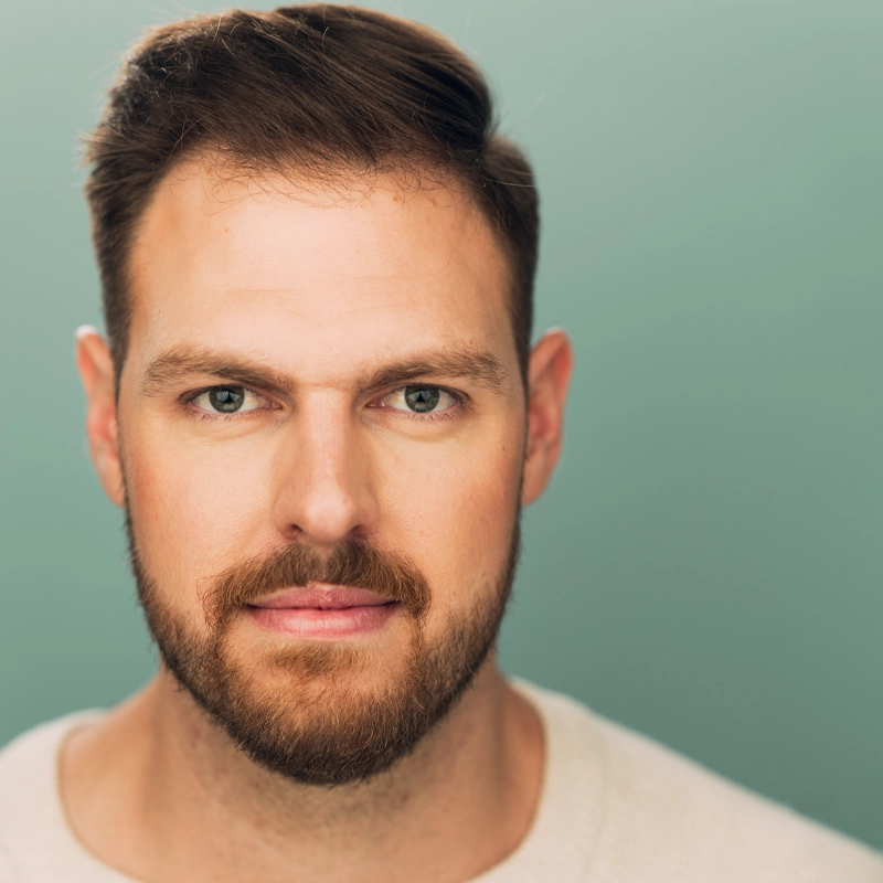 A man with short brown hair, a neatly trimmed beard, and mustache looks directly at the camera. He wears a light-colored crew-neck sweater. The background is a solid, muted teal color. The lighting is soft, highlighting his facial features.