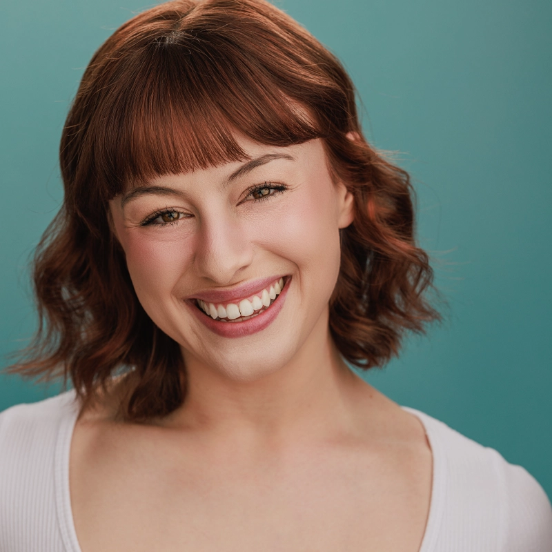 A young woman with wavy, shoulder-length auburn hair and bangs smiles warmly. She has fair skin, subtle makeup, and is wearing a white top. The background is a solid teal color, and the lighting is bright and even.