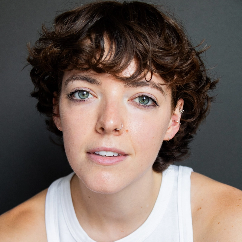 A person with short, wavy brown hair, light skin, and green eyes looks at the camera. They wear a white sleeveless top and two nose rings, with small earrings visible. The background is plain and dark, highlighting the person’s face.