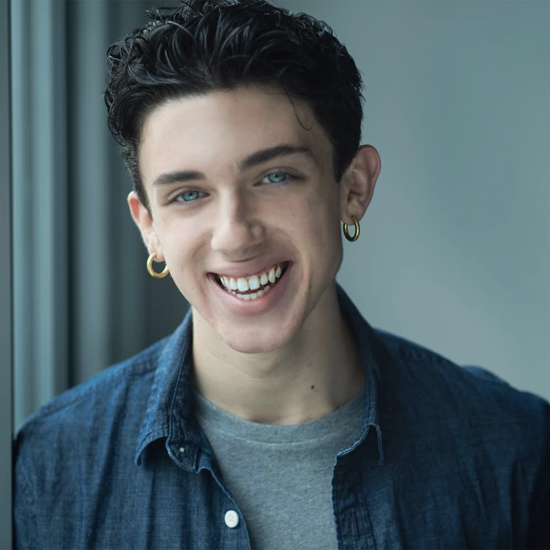 A young person with short, dark curly hair and blue eyes smiles warmly. They wear gold hoop earrings, a gray t-shirt, and a blue button-up shirt. The background is softly blurred, suggesting a window or indoor setting with natural light.