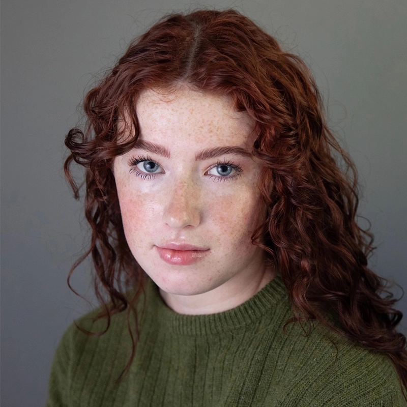 Isabella Greathouse, a young woman with fair skin, blue eyes, and long, curly red hair, looks at the camera. She has light freckles and wears a green ribbed sweater. The background is plain gray with soft, even lighting.