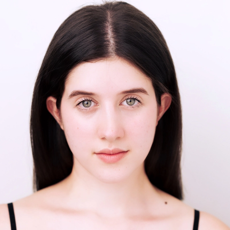 A young woman with long, straight dark hair and fair skin stares directly at the camera. She has light eyes, soft pink lips, and is wearing a black strap top. The background is plain and white, giving the portrait a clean, minimalist look.