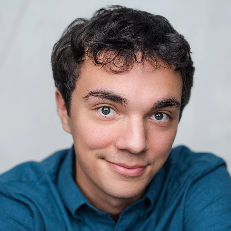 A young man with short, dark hair and light skin smiles gently at the camera. Michael Mariniello is wearing a blue button-up shirt and is set against a light, plain background. The image is cropped closely on his face and shoulders.