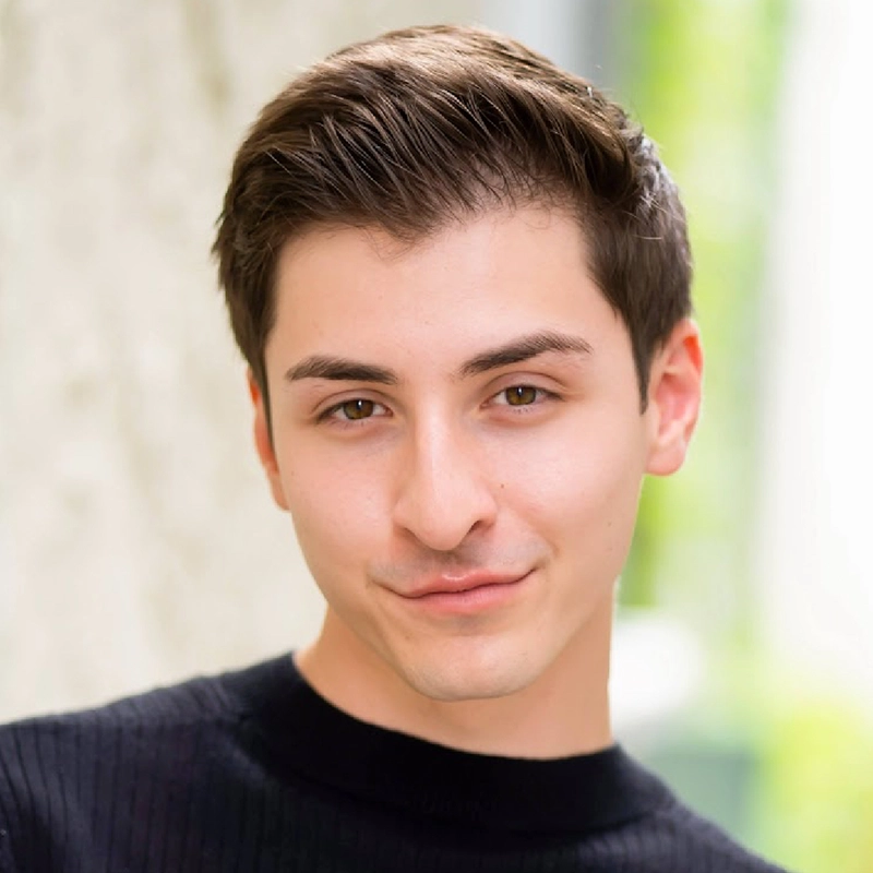 RJ Vandenbrouck, a young man with short brown hair and fair skin, is smiling subtly. He wears a black ribbed sweater and stands before a softly blurred outdoor backdrop with hints of greenery and gentle, natural lighting.