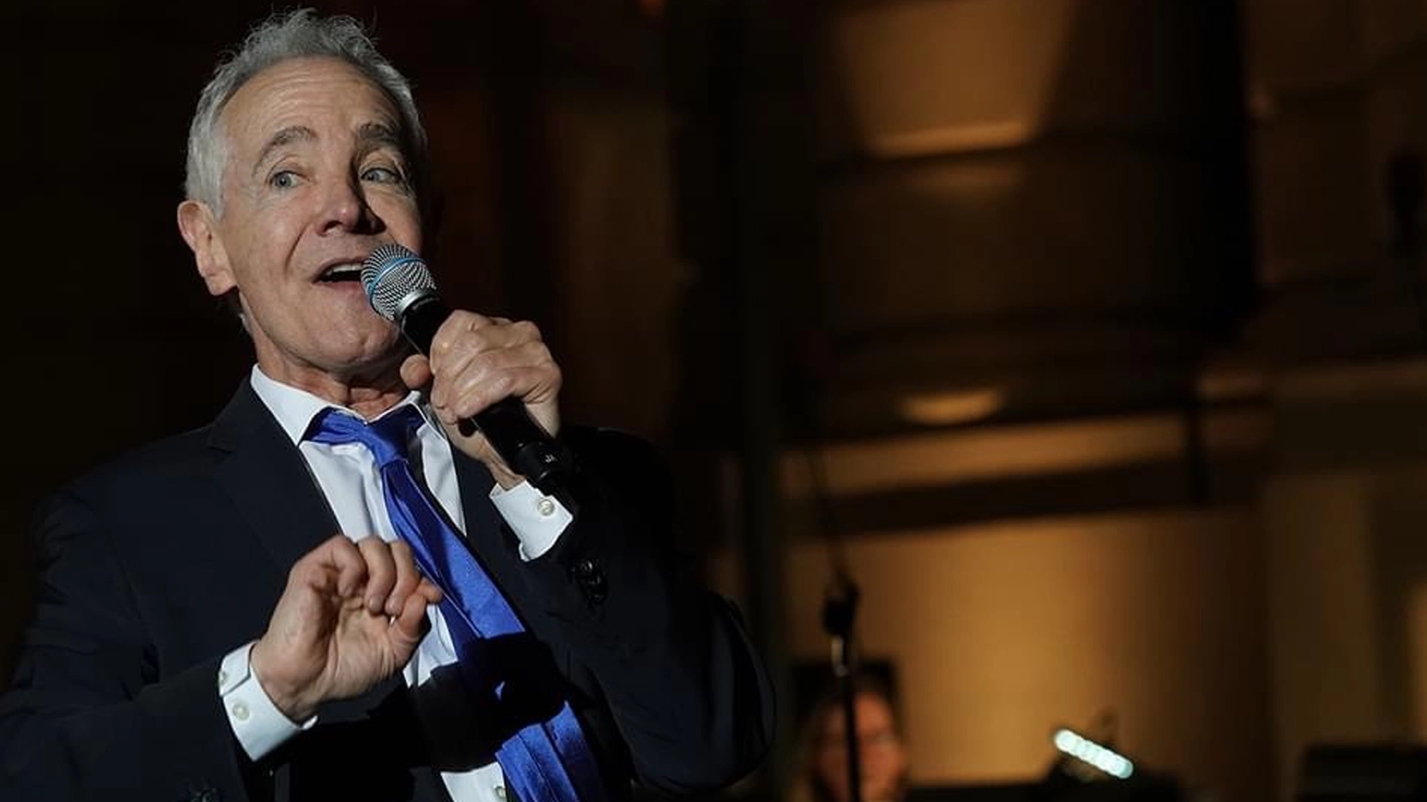 An older man in a suit and blue tie holds a microphone and sings on stage, reminiscent of Jason Graae. He gestures with his left hand under the night’s dim lighting, shadows cast behind him to emphasize the performer.