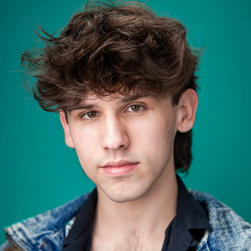 Caroline Weston, a young person with wavy brown hair and fair skin, looks directly at the camera. Wearing a blue denim jacket over a dark shirt, she stands against a teal green background, her expression neutral and calm.