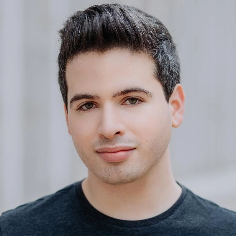 A young man with short, dark hair and fair skin, wearing a black crew-neck shirt, looks directly at the camera with a neutral expression. The softly blurred, light gray background evokes the portrait style of Caroline Weston.