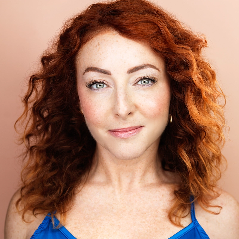Jenna Coker-Jones, a woman with curly red hair and fair, freckled skin, smiles softly at the camera. She has light-colored eyes, subtle makeup, and wears a blue top against a solid peach background, creating a warm, inviting portrait.