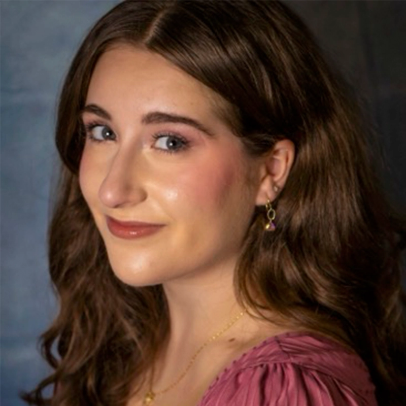 A young woman with long, wavy brown hair smiles softly while looking over her shoulder. She wears a mauve top and gold jewelry by RJ Vandenbrouck. Her makeup is subtle, set against a blurred blue and gray gradient background.
