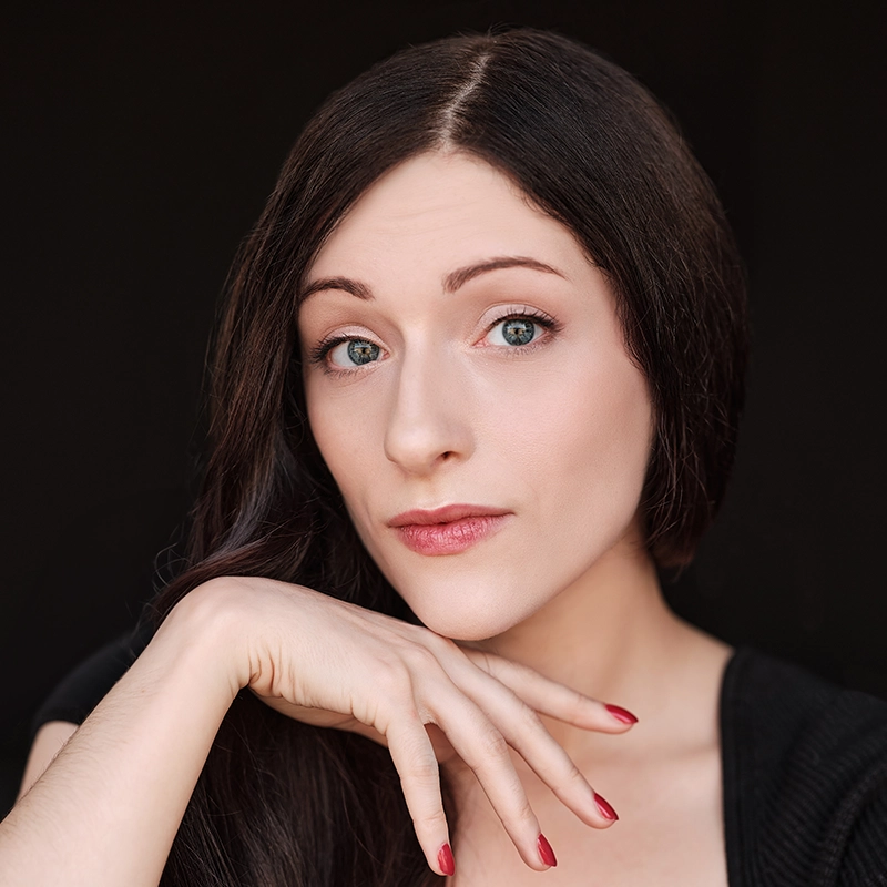 Sahara Williams, with long, straight dark hair and fair skin, looks at the camera with a neutral expression. She rests her chin on her hand, red-painted nails visible. Wearing a black top, she stands out against the dark background.