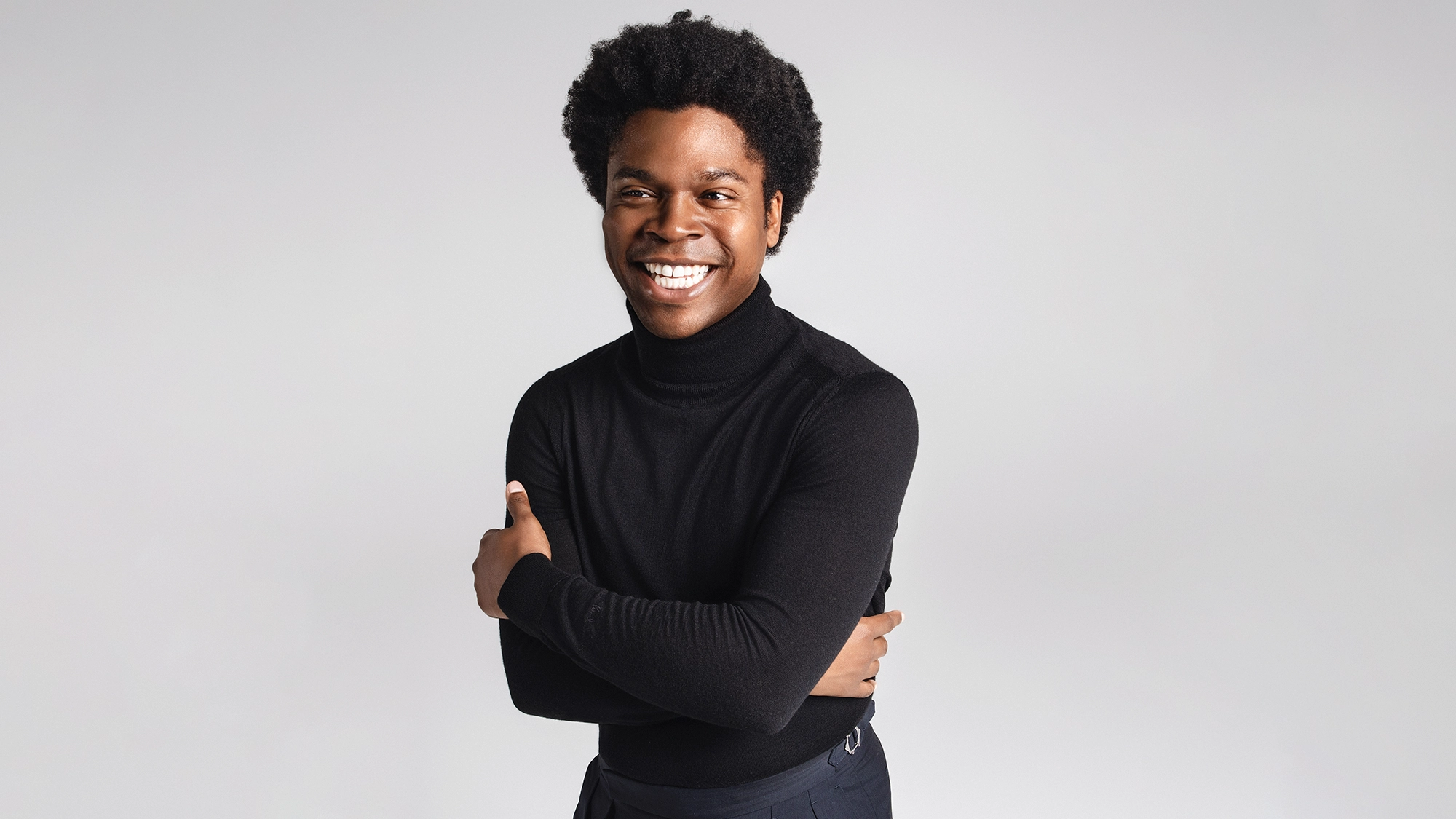 A smiling singer with short curly hair stands against a light gray background, wearing a black turtleneck and dark pants. Their arms are crossed comfortably, and their expression is friendly and approachable—just like Jimmie Herrod of America’s Got Talent.