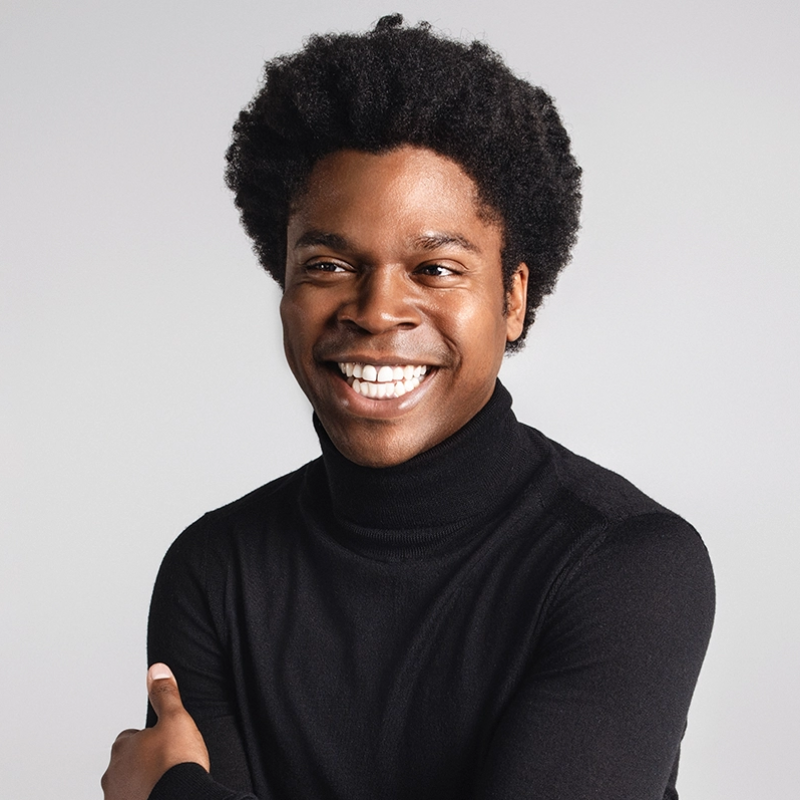 A young man with dark skin and short curly hair smiles broadly, showing his teeth. He is wearing a black turtleneck sweater with his arms crossed, posed against a plain light grey background. The photo conveys a cheerful and friendly mood.