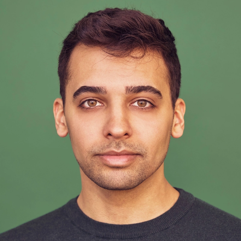 Peyton Amaral, a young man with short dark hair and hazel eyes, looks directly at the camera with a neutral expression. He has trimmed facial hair, wears a dark crew-neck sweater, and stands against a solid green background.