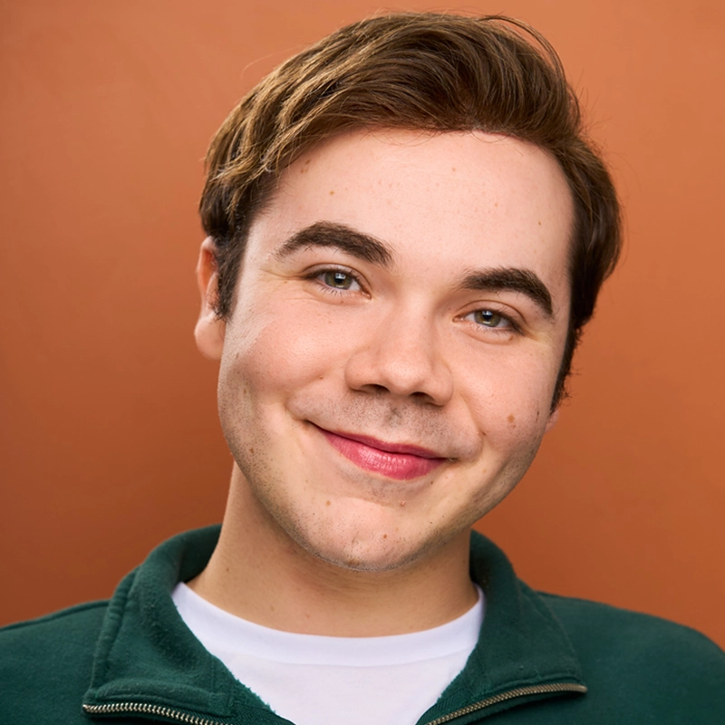 Young adult Michael Mariniello, with short brown hair, is wearing a green zip-up jacket over a white shirt, smiling warmly at the camera. The background is solid orange. The lighting is bright and even, highlighting Michael’s friendly expression.