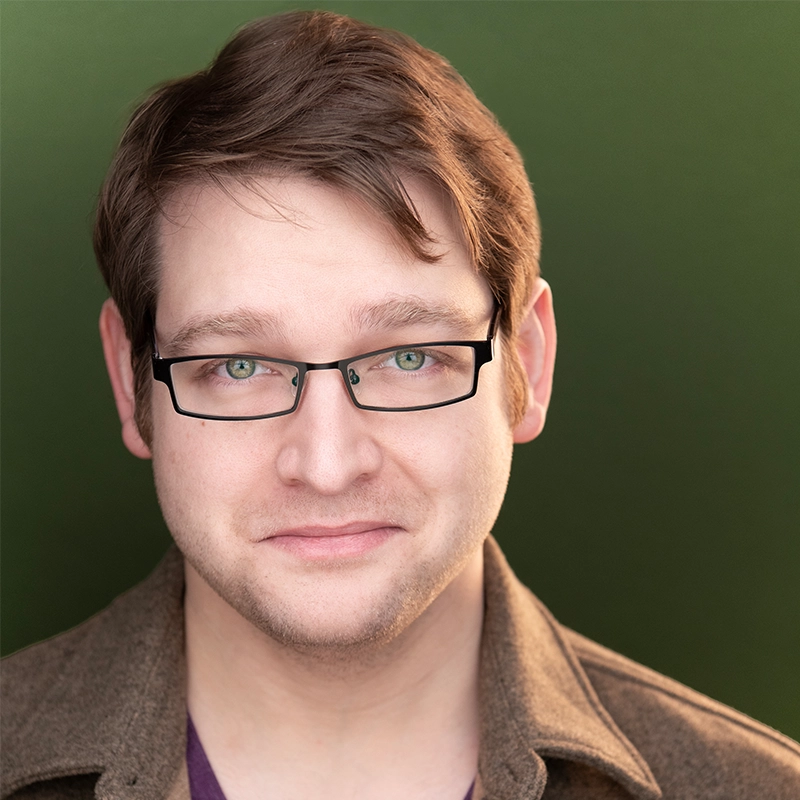 Lauren Torey, with fair skin, short light brown hair, and light-colored eyes behind rectangular black glasses, smiles softly. Dressed in a brown jacket over a purple shirt, she stands against a blurred dark green background.