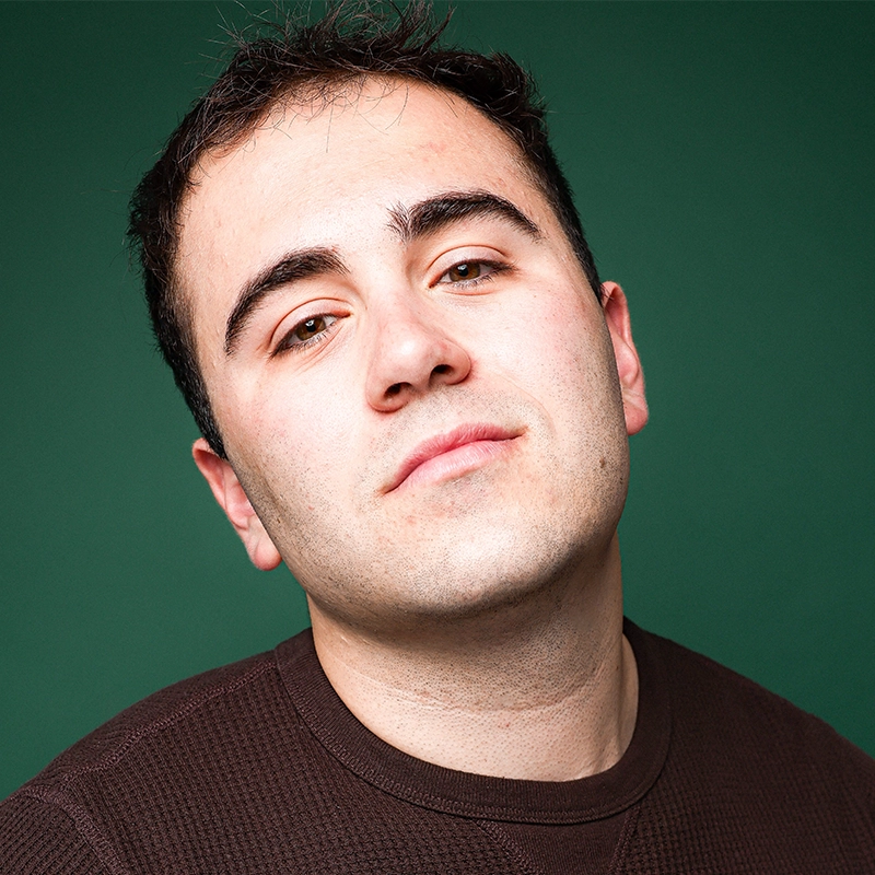 A young man with short dark hair and pale skin tilts his head slightly and looks confidently at the camera. He wears a dark brown textured shirt. The background is a solid dark green, in the distinctive style of Lauren Torey. Even lighting highlights his features.