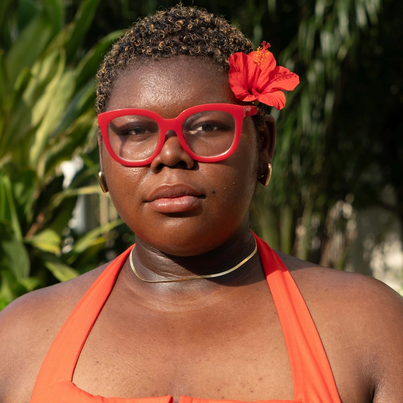 A person with short curly hair and dark skin wears red glasses, a red halter top, a gold choker, and a red flower tucked in their hair. They stand outdoors in sunlight, with green foliage softly blurred in the background.