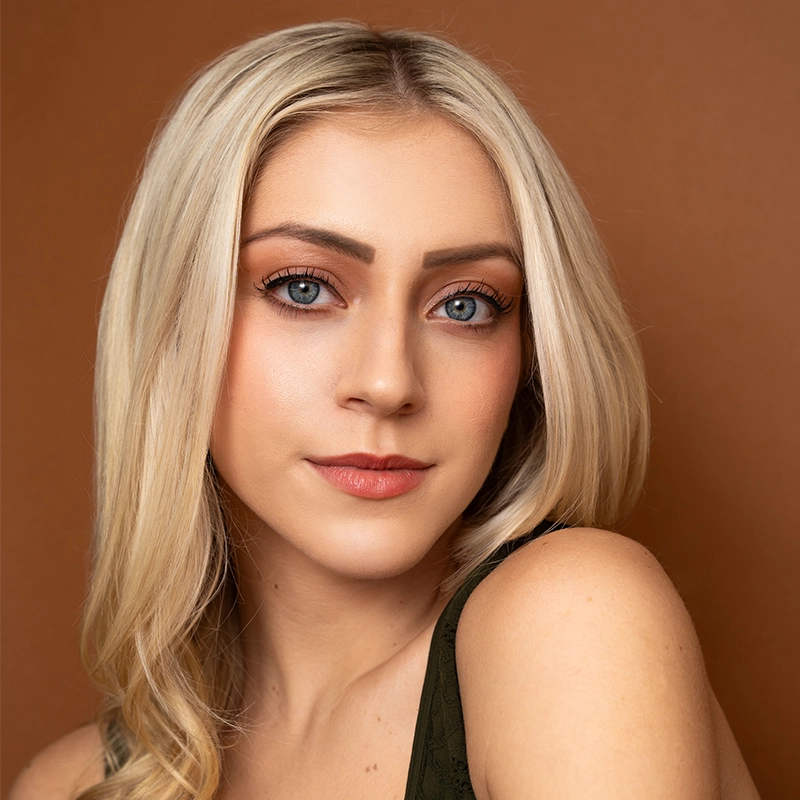 Peyton Amaral, a young woman with long, straight blonde hair and blue eyes, poses in front of a brown background. She wears natural makeup with defined eyebrows and nude lipstick, exuding a soft, confident expression.