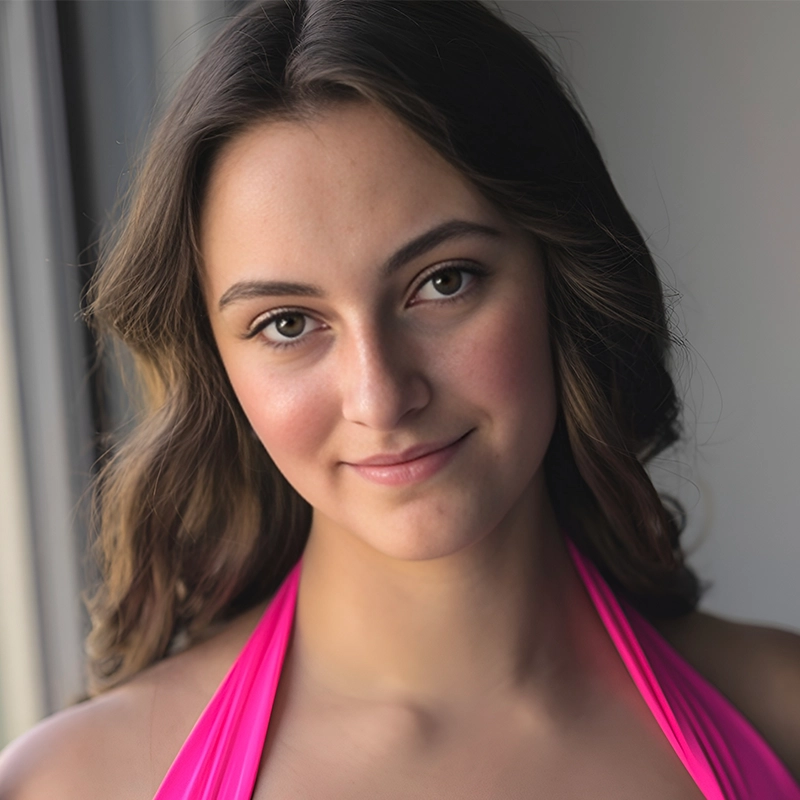Danielle Serrano-Bremer, a young woman with wavy brown hair and light skin, smiles softly in a bright pink halter top. She stands near a window as soft natural light illuminates her face against a neutral, slightly blurred background.