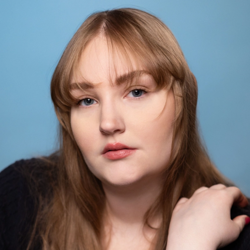 Caroline Weston, a young woman with long light brown hair and bangs, poses against a light blue background. She has fair skin, blue eyes, and natural makeup, looking calmly at the camera with her hand resting on her shoulder.