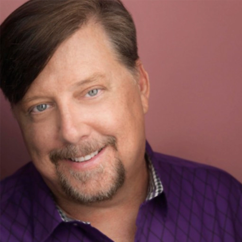 A smiling middle-aged man with light skin, blue eyes, and a trimmed goatee wears a purple checkered shirt. His short brown hair is neatly side-parted. The background is a solid muted mauve, in the distinctive portrait style of Caroline Weston.