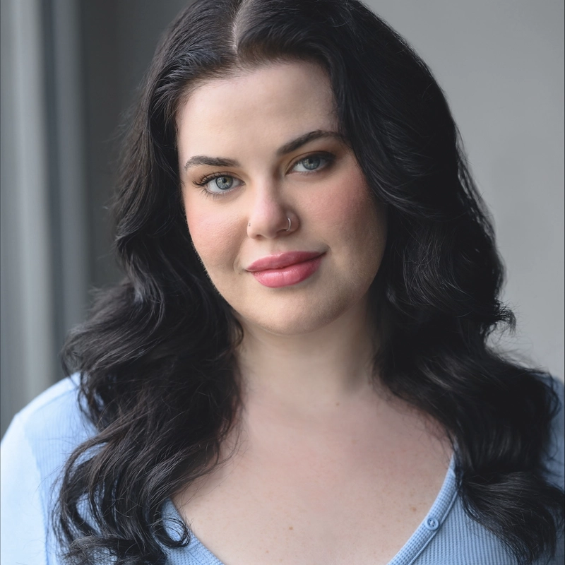 A woman with long, wavy dark hair smiles softly. She has light skin, blue eyes, and wears natural makeup with pink lipstick. Sporting a nose ring and a light blue v-neck top, she stands before a softly blurred gray background—photographed by Ricky Kessler.