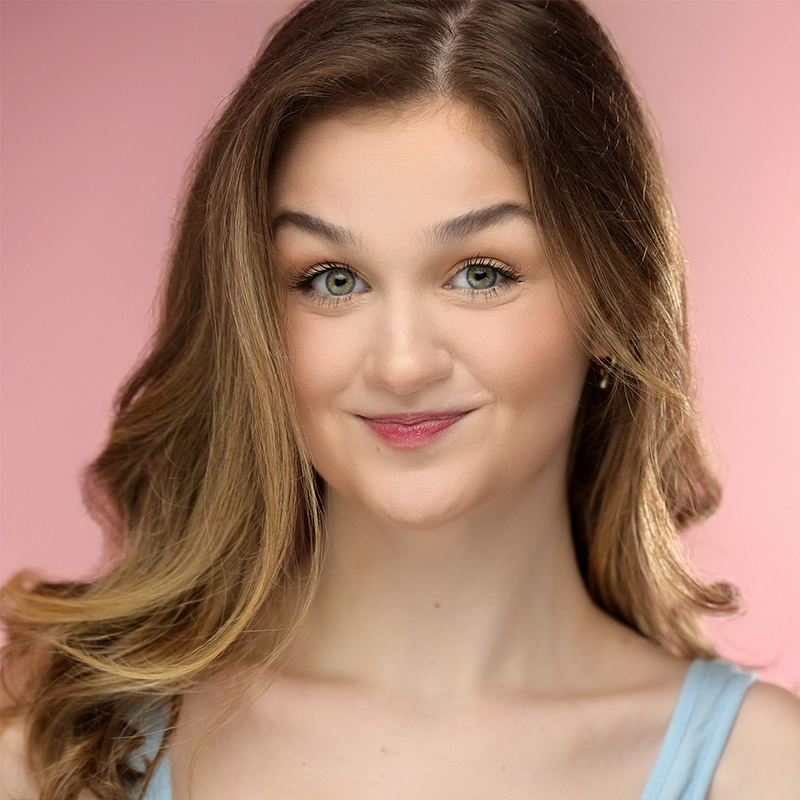 Sahara Williams, a young woman with long, wavy light brown hair and green eyes, smiles slightly. She wears a light blue tank top and subtle makeup. The background is a soft pink gradient with bright, even lighting.