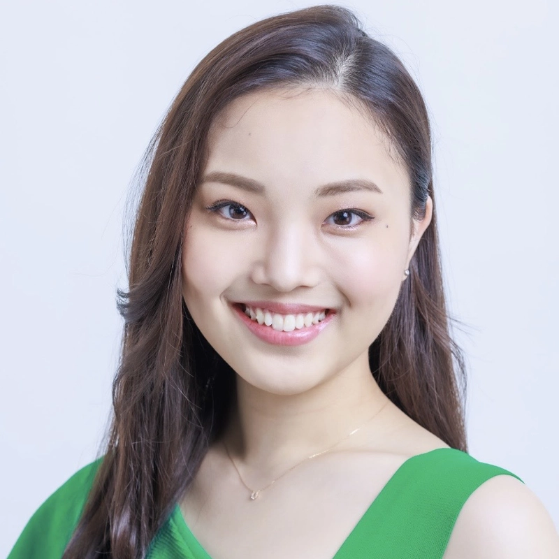 A young woman with long, straight brown hair and fair skin smiles warmly at the camera. She wears a bright green top and a delicate gold necklace. The background is a plain, light blue, drawing attention to her friendly expression.