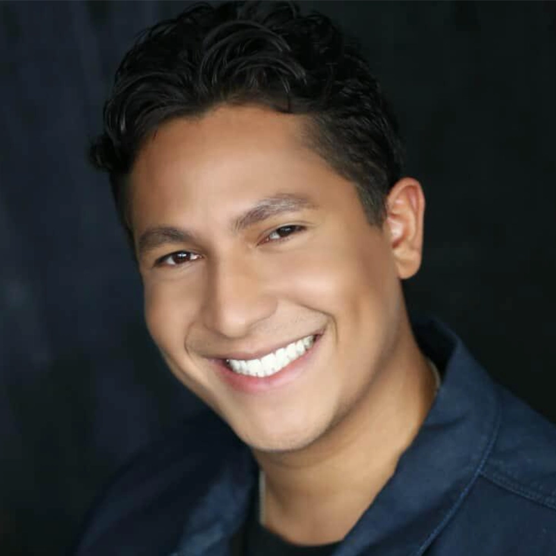 A smiling man with short, dark, wavy hair wears a dark blue jacket over a black shirt and poses in front of a dark, blurred background. Even lighting highlights his face as he looks directly at the camera, in the signature style of Caroline Weston.