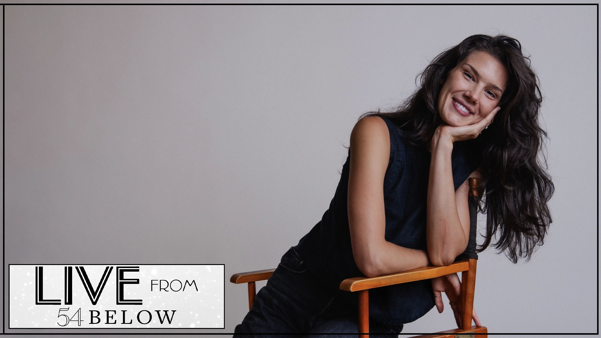 LIVE Christine Oczkus A woman with long dark hair, wearing a sleeveless dark top, sits sideways on a wooden chair, smiling and resting her chin on her hand. The background is plain light gray. In the bottom left corner, “LIVE FROM 54 BELOW” promotes the livestream of Christine Oczkus.