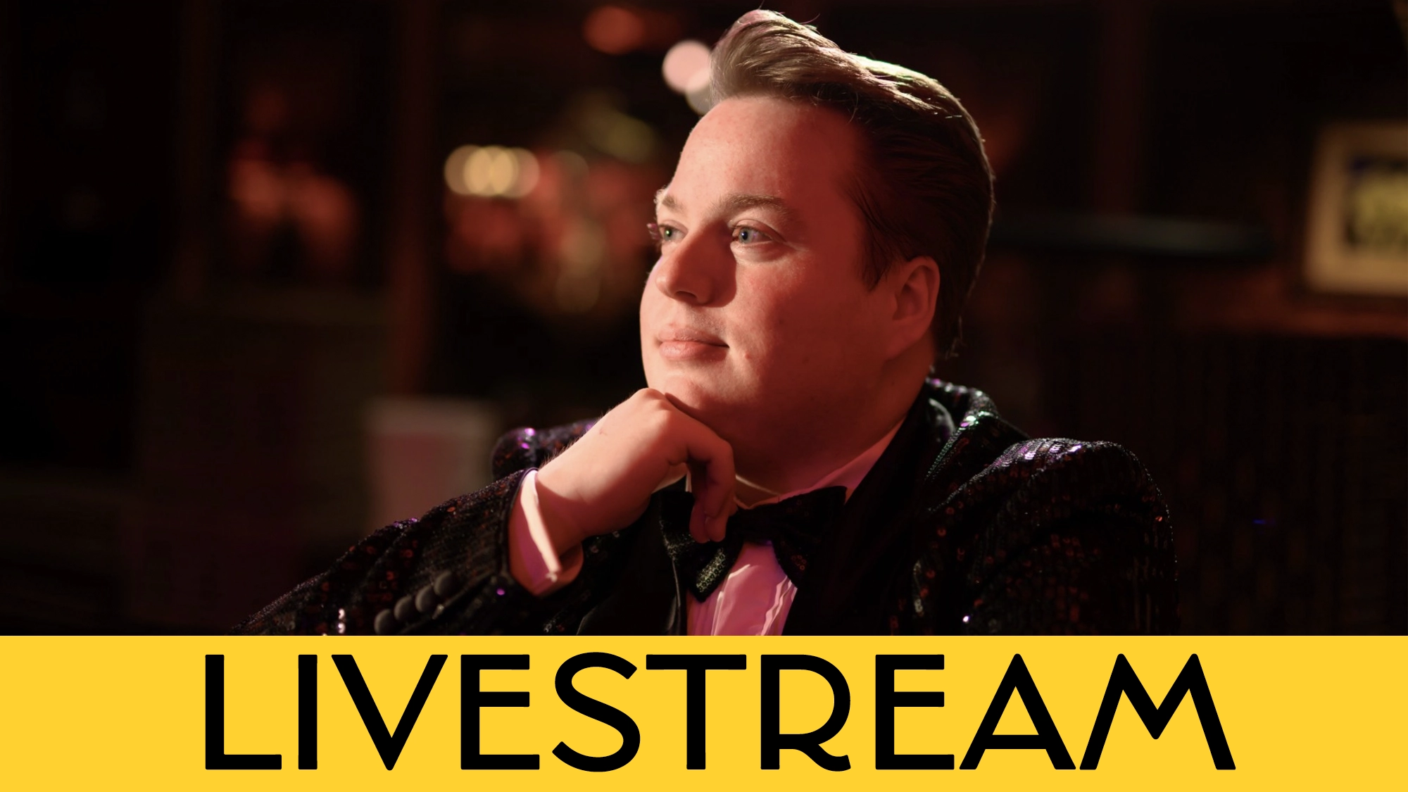 A person in a tuxedo with a sequined jacket sits thoughtfully with their chin resting on their hand. The background is softly blurred with warm lighting. A bold yellow banner reads “LIVESTREAM: MACON Prickett—His Own Way” in black letters.