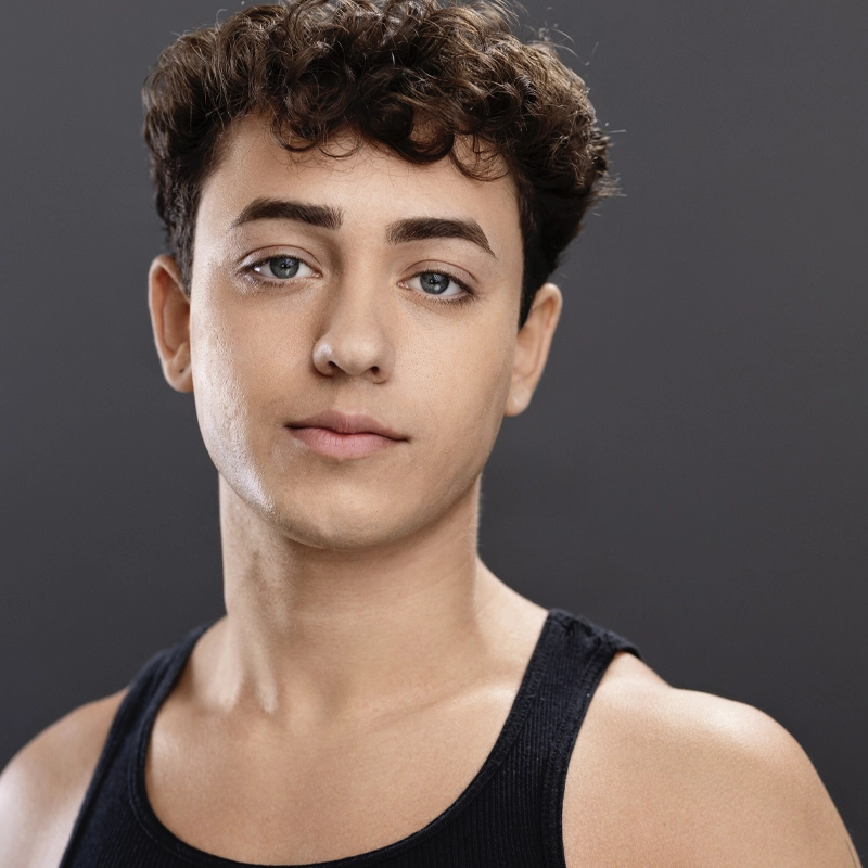 A young person with short, curly brown hair and blue eyes looks at the camera with a neutral expression. Wearing a black tank top, Andre Lajevardi stands against a solid dark gray background, the even lighting highlighting their face and shoulders.