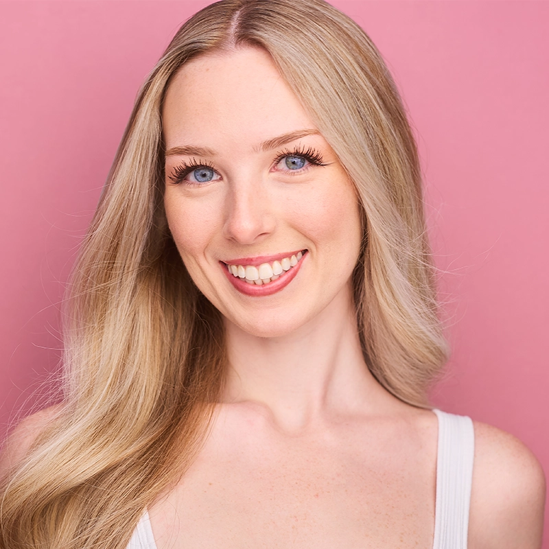 Caroline Bowers, a young woman with long, straight blonde hair, smiles at the camera. She wears a white tank top and has natural makeup with long eyelashes. The solid pink background highlights her tilted head and hair draped over one shoulder.