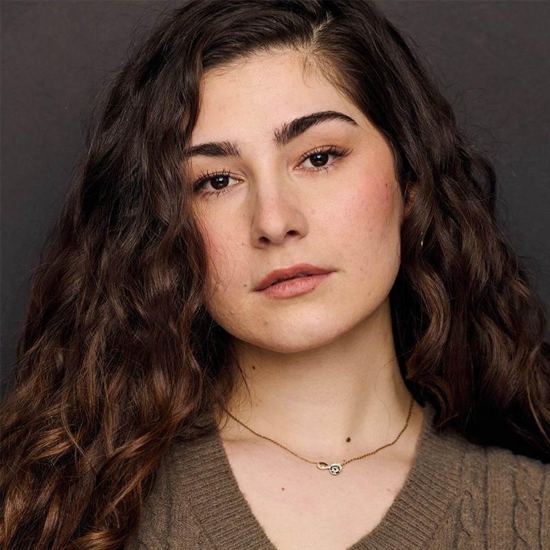Laura Pachnos, a young woman with long, wavy brown hair and bushy eyebrows, poses against a dark background. She wears a brown knit sweater and a delicate gold necklace with a small pendant, gazing neutrally into the camera.