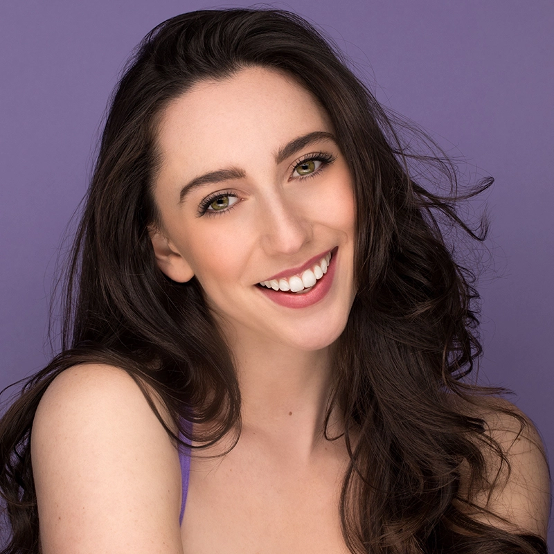 A young woman with long, wavy brown hair smiles warmly at the camera. She has light skin, hazel eyes, and wears natural makeup. Dressed in a purple top, Mickey Wirtz is posed against a solid lavender background.