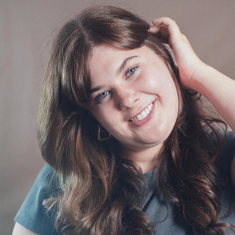 Caroline Bowers, a woman with long brown hair and blue eyes, smiles warmly. She wears a dark top and gold hoop earrings, with one hand touching her hair. The plain, softly lit background highlights her friendly expression.