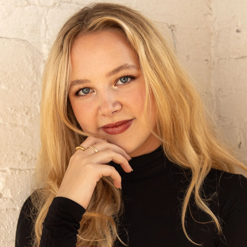 Laura Pachnos, a young woman with long blonde hair and blue eyes, poses in front of a textured white brick wall. She wears a black turtleneck and gold rings, resting her chin on her hand and smiling softly with relaxed confidence.