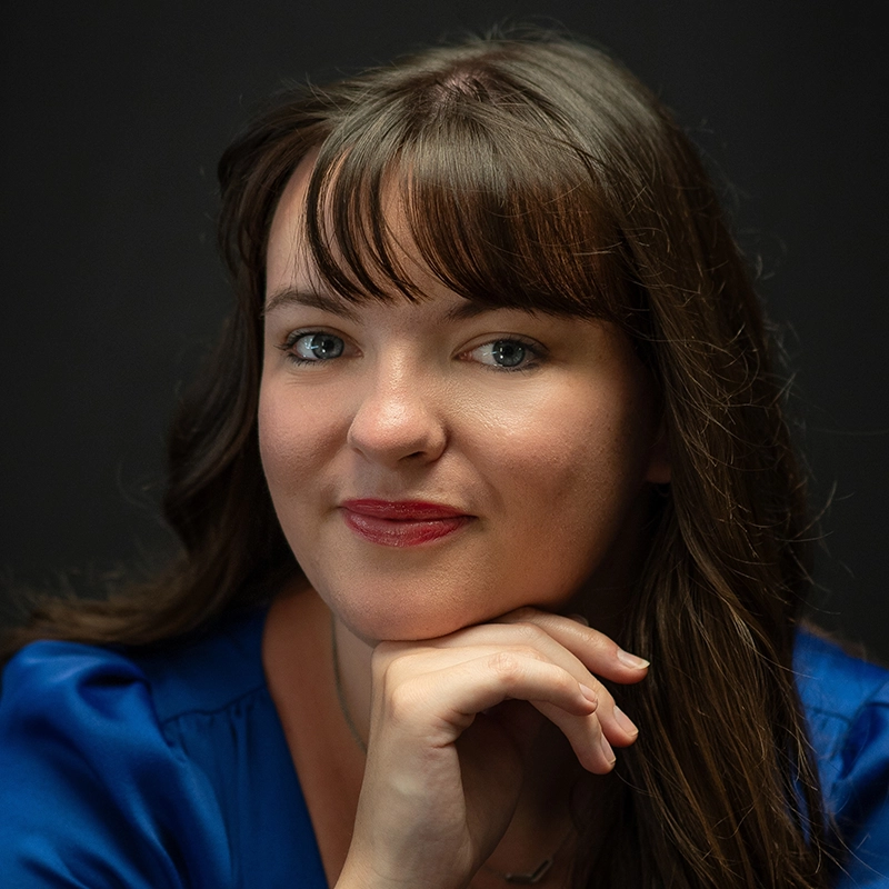 Lauren Torey, a woman with long brown hair and bangs wearing a blue top, poses in front of a dark background. She smiles slightly, resting her chin on her hand and looking directly at the camera with a calm expression.