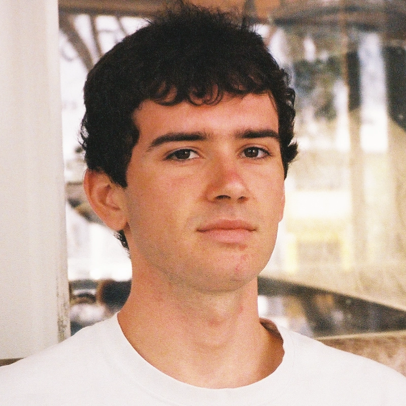 A young man with short, dark curly hair and fair skin, Daniel Legare, looks slightly past the camera with a neutral expression. He is wearing a plain white shirt. The blurred background features windows and subtle indoor reflections.